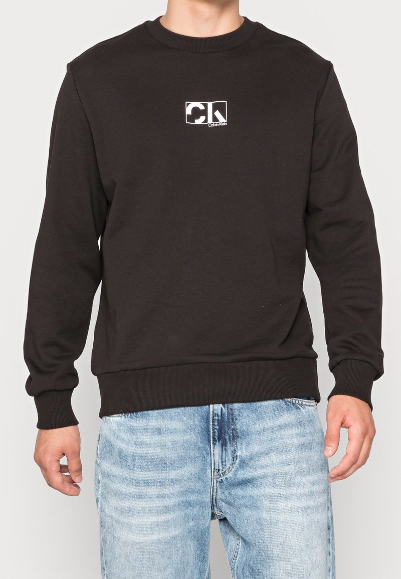 Homme portant un sweat Calvin Klein noir avec un petit logo blanc sur la poitrine et un jean bleu clair, debout devant un fond uni.