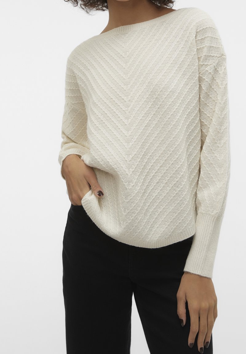 Femme portant un pull en maille crème avec un motif en chevrons et un pantalon noir, main dans la poche, sur fond blanc.