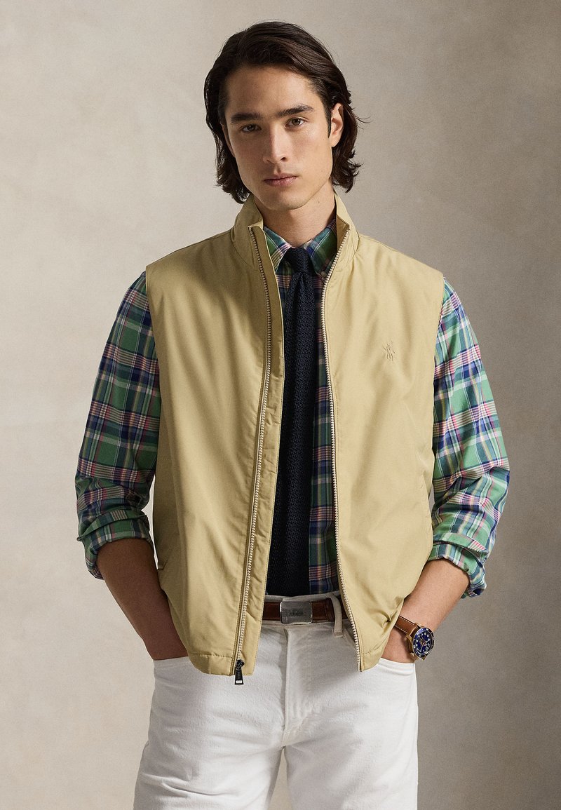 Μπεζ μπουφάν χωρίς μανίκια πάνω απ ό πουκάμισο σε πράσινο και μπλε καρό. Λευκό παντελόνι και σκούρα γραβάτα. Σημαντικά χαρακτηριστικά περιλαμβάνουν φερμουάρ και πλαϊνές τσέπες.