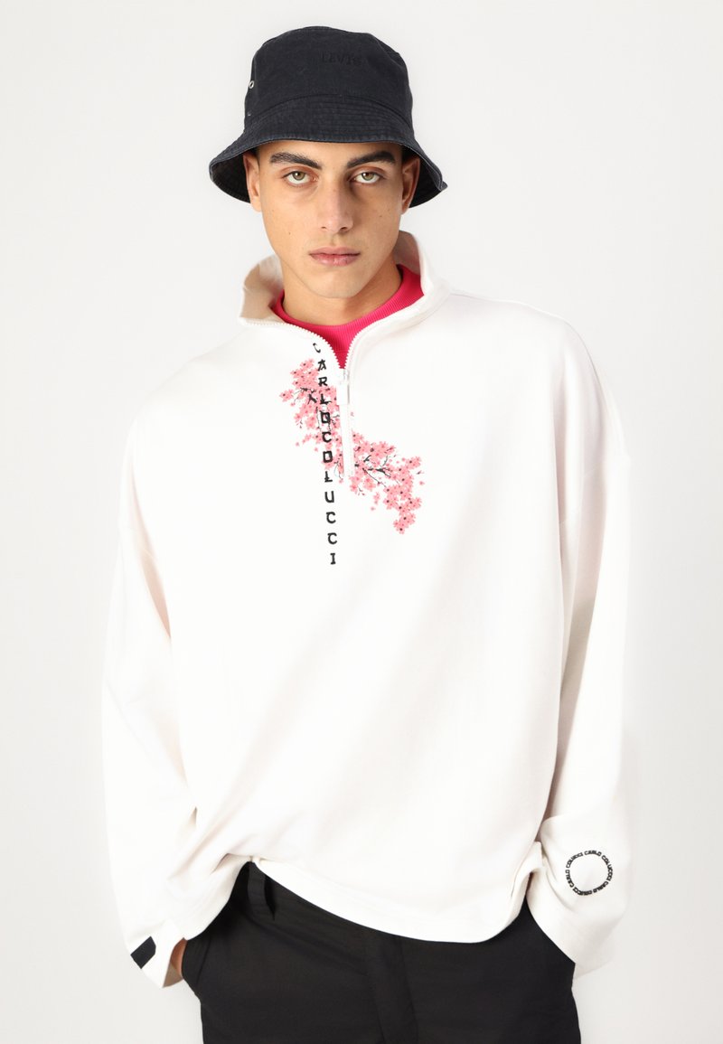 Biały polarowy pulower z suwakiem, ozdobiony różowym haftem kwiatowym i czarnym napisem, uzupełniony granatowym kapeluszem typu bucket.