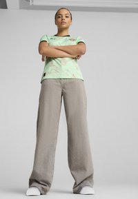 Haut vert à manches courtes avec un motif texturé et des accents noirs, associé à un pantalon large gris et des baskets blanches, vu d'en bas.