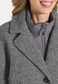 Femme souriante portant un manteau en laine gris texturé par-dessus une veste rembourrée grise zippée avec col, montrant la partie supérieure de la poitrine et le cou.
