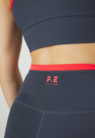 Jūrinė trumpa palaidinė ir suderinti aukšti tamprės su raudonomis detalėmis. Juosmens vietoje yra raudonas logotipas "P.E NATION". Lygi tekstūra.