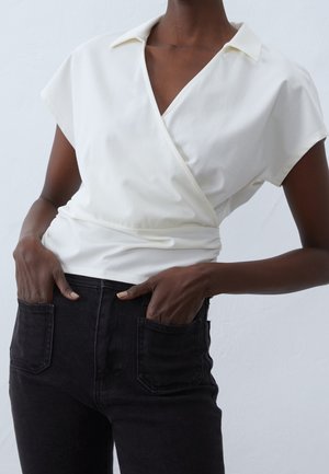 Witte overslagtop met kraag, korte mouwen en een getailleerde taille. Gecombineerd met hooggetailleerde zwarte spijkerbroek met voorzakken.