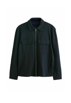 Person, der spænder manchetterne på en mørk marineblå jakke med en synlig lynlås foran, tæt på hænder og stoftekstur.