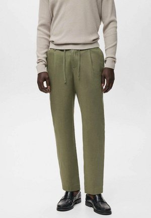 Pantalones verde olivo con pliegues, cintura con cordón y corte a la altura del tobillo. Combinados con un suéter beige de canalé y zapatos negros.