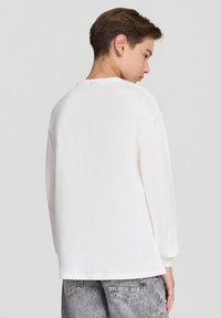 Biała koszula z długim rękawem o okrągłym dekolcie, wykonana z miękkiego materiału, o luźnym kroju i zaokrąglonym dolnym wykończeniu, w połączeniu z szarymi dżinsami.