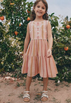 Joven niña con un vestido rosa a rayas y sandalias blancas está sonriendo al aire libre cerca de un naranjo con fruta madura.