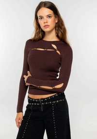 Top largo de manga larga marrón con relieve, que presenta detalles recortados acentuados con botones decorativos. Combinado con pantalones negros adornados con tachuelas.