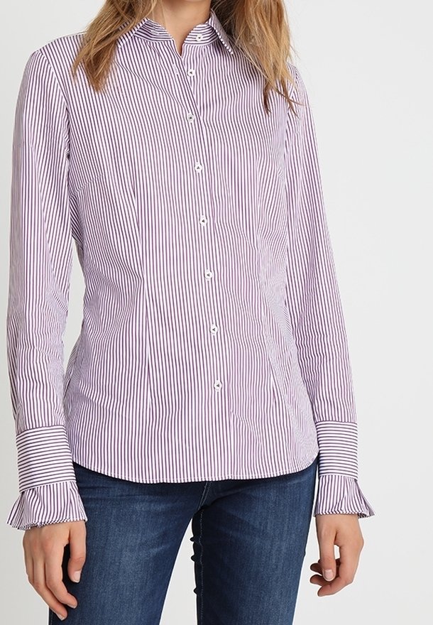 Femme portant une chemise blanche ajustée à manches longues avec de fines rayures verticales violettes et des poignets froufroutants, associée à un jean bleu foncé.