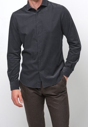 Mężczyzna w ciemnoszarej koszuli zapinanej na guziki z długimi rękawami i brązowych spodniach, stojący z jedną ręką w kieszeni, na jednolitym tle.
