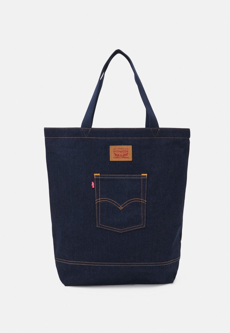 Sac fourre-tout en denim bleu foncé avec poche avant, écusson de marque en cuir et double poignée pour le transport.