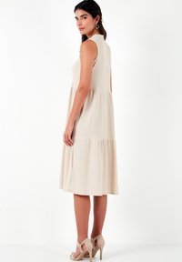 Beige sleeveless dress with a tiered design and textured fabric, featuring a high neckline and side cutouts. Worn with beige heeled sandals.