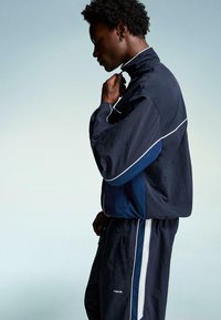 Marine tracksuit med kontrasterende hvide og blå detaljer. Har en høj krave og en løs pasform. Stoffet synes let og struktureret.