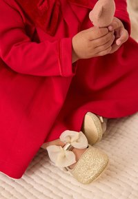 Kuldse sädelusega baleriinikingad beežikaunistusega ning erkpunase pika varrukaga kleidiga, paigutatud tekstuurse valge pinna peale.