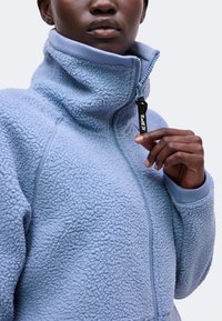 Personne portant une veste polaire texturée bleu clair, tenant le curseur de la fermeture éclair près du col avec une étiquette en tissu noire attachée.