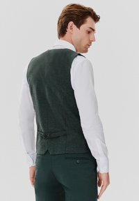 Ciemnozielona kamizelka wykonana z teksturowanego materiału, charakteryzująca się dopasowanym krojem, kieszeniami z tyłu i dwoma guzikami do regulacji, noszona na białej koszuli.