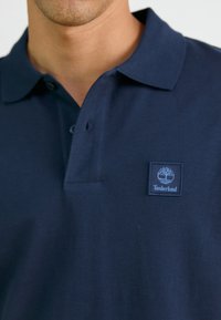 Jūras spēku krāsas polo krekls, izgatavots no kokvilnas, ar trīs pogu aizdari, īsām piedurknēm un Timberland logu uz kreisā krūtīm.