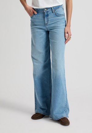 Kobieta ubrana w jasnoniebieskie dżinsy z wysokim stanem i szerokimi nogawkami, biały top, brązowe mokasyny oraz złote pierścionki na prawej ręce.