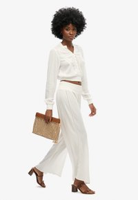 Blusa blanca corta con intrincados detalles en el frente, combinada con pantalones de pierna ancha de tiro alto a juego. Sosteniendo un bolso de mano tejido en color marrón claro.
