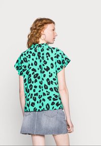 Top verde com padrão de leopardos, com mangas curtas e esvoaçantes, fecho com zíper nas costas, combinado com uma saia mini de denim cinza.
