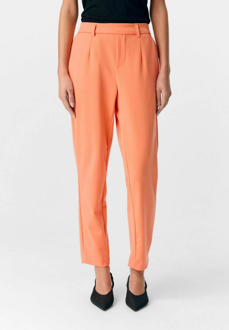 Pantalons couleur pêche avec une coupe ajustée, dotés d'une taille haute, de plis à l'avant et de jambes fuselées. Associés à des escarpins noirs à bout pointu.