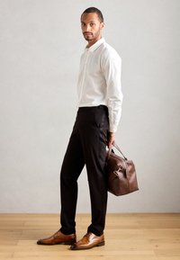 Camisa blanca de botones, pantalones negros, zapatos de cuero marrón y un bolso de cuero marrón. Cabello corto, complexión media, de pie sobre un suelo de madera.
