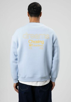 Lichtblauwe sweatshirt met gele tekst op de achterkant: "dromen najagen, NIET deadlines." Heeft een losse pasvorm en geribbelde manchetten.