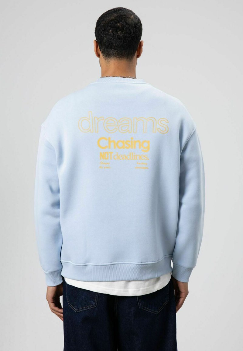 Sweat-shirt bleu clair avec texte jaune au dos : « dreams Chasing NOT deadlines. » Présente une coupe décontractée et des poignets côtelés.