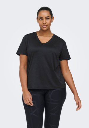 Kvinde iført en ensfarvet sort V-hals T-shirt og sorte leggings med subtile abstrakte mønstre, stående mod en lysegrå baggrund.