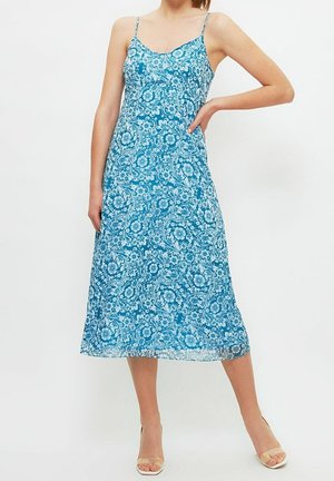 Kobieta w niebieskiej, kwiatowej sukience midi na cienkich ramiączkach i beżowych sandałkach na obcasie, pozująca z jedną ręką na biodrze na białym tle.