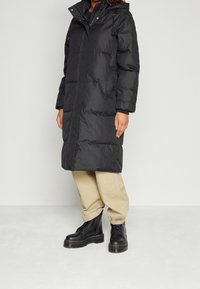 Černý péřový kabát s kapucí, texturovaná látka, dlouhý střih, přední zip, boční kapsy, nošený přes béžové kalhoty a černé šněrovací boty.