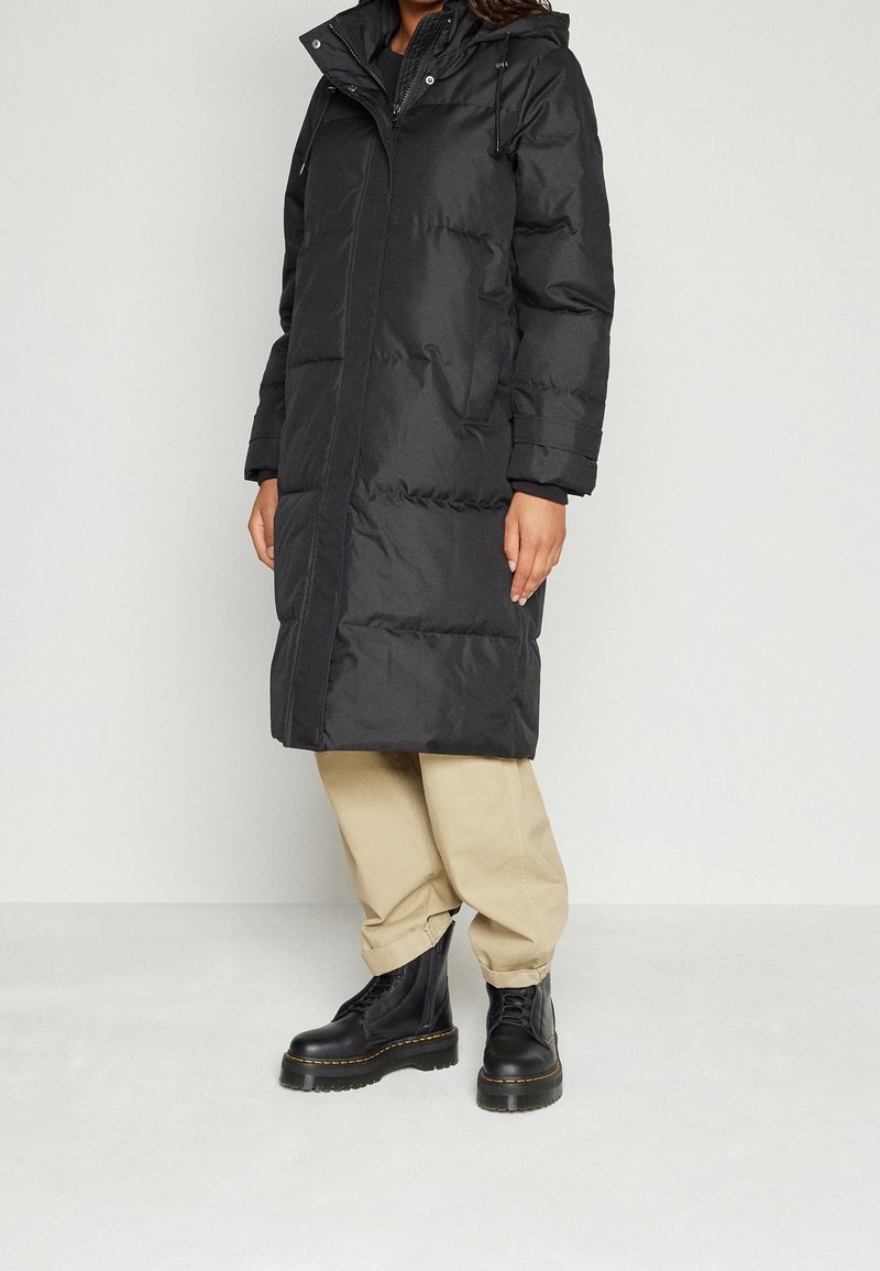 Černý péřový kabát s kapucí, texturovaná látka, dlouhý střih, přední zip, boční kapsy, nošený přes béžové kalhoty a černé šněrovací boty.