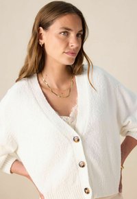 Jonge vrouw met lichtbruin haar, draagt gouden creolen, gelaagde gouden kettingen en een witte geknoopte cardigan over een kanten top.