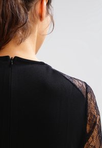 Černé šaty s hladkým tělem z látky a krajkovými rukávy, s klíčovou dírkou na zádech a kulatým výstřihem.