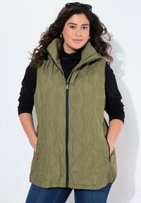 Gilet trapuntato verde oliva con colletto alto, zip completa e tasche laterali, indossato sopra un top a maniche lunghe nero e abbinato a jeans blu.