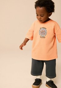 Lichtoranje t-shirt met een ronde afbeelding met tekst. Gecombineerd met donkergrijze shorts en zwarte sneakers, witte sokken zichtbaar.