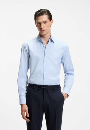 Mężczyzna w jasnoniebieskiej koszuli w małą kratkę i ciemnoniebieskich spodniach ze ściągaczem, stoi z jedną ręką w kieszeni na białym tle.