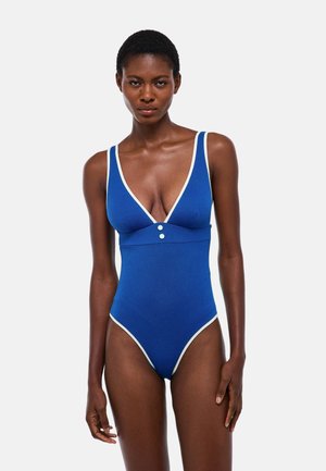 Femme portant un maillot de bain une-pièce bleu avec bordure blanche et boutons, debout devant un fond clair uni.
