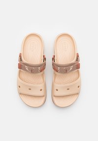 Beiga Crocs-sandaler med justerbara tygremmar som har ett brunt och orange mönster, samt ventilerat tåområde, visas uppifrån.