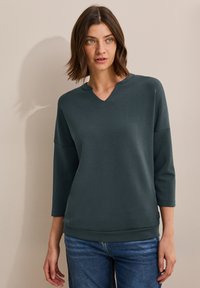 Suéter verde oscuro con cuello en V, mangas de tres cuartos y corte relajado. Hecho de una tela suave, presenta un dobladillo y puños acanalados.