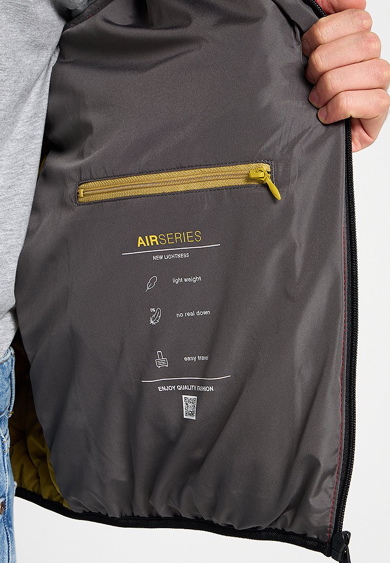 Interno grigio di una giacca con una tasca gialla con zip. Etichette stampate indicano design leggero, senza piumino reale e facilità di viaggio.