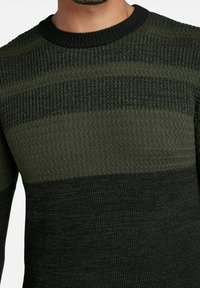 Sweter dzianinowy w ciemnozielonym i oliwkowym kolorze, z teksturowanymi paskami i prążkowanym dekoltem w serek. Miękka tkanina z różnorodnymi wzorami szycia.