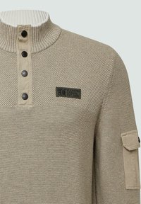 Beżowy sweter dzianinowy z prążkowanym wysokim kołnierzem, czterema czarnymi zatrzaskami, kieszenią na rękawie oraz etykietą "s.O Elevated Casual Est. 1969".