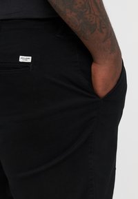 Zwarte denim broeken met een zijzak en een klein wit merklabel. De stof heeft een gladde textuur en een getailleerd ontwerp.