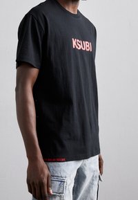 Černé bavlněné tričko s růžovým logem "KSUBI" na přední straně a červeným textem "NĚKTEŘÍ MI VĚŘÍ" na lemu. Klasický střih s kulatým výstřihem.