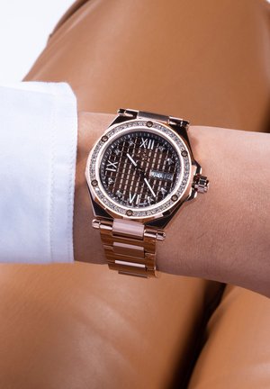 Rose gold watch with octagonal case, brown patterned dial, Roman numerals, diamond accents, and a linked strap on a wrist.