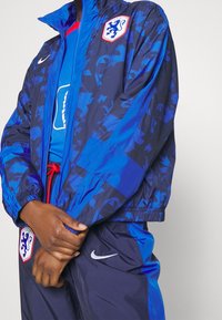 Blåmönstrad vindjacka med frontdragkedja, röd kontrastdragkedja och logotypmärken. Kombinerad med en blå sporttopp under.