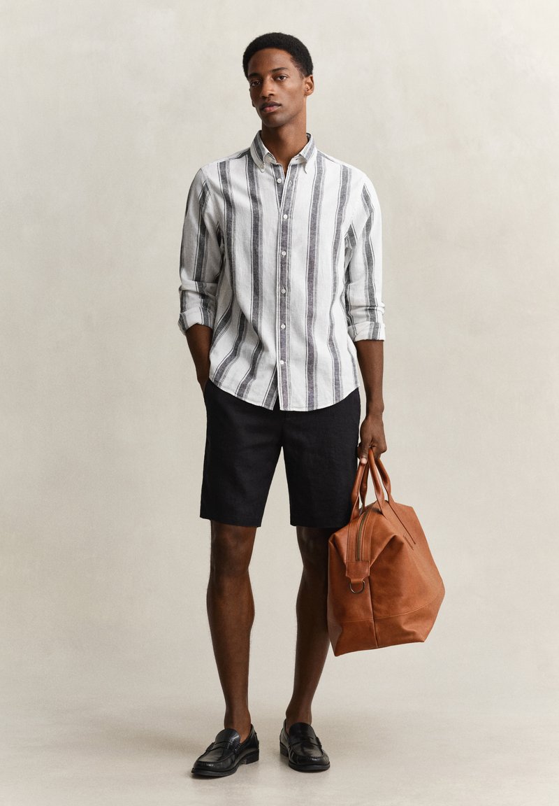 Man die een wit en grijs verticaal gestreept overhemd draagt met zwarte shorts en zwarte loafers, en in één hand een grote bruine leren tas vasthoudt.