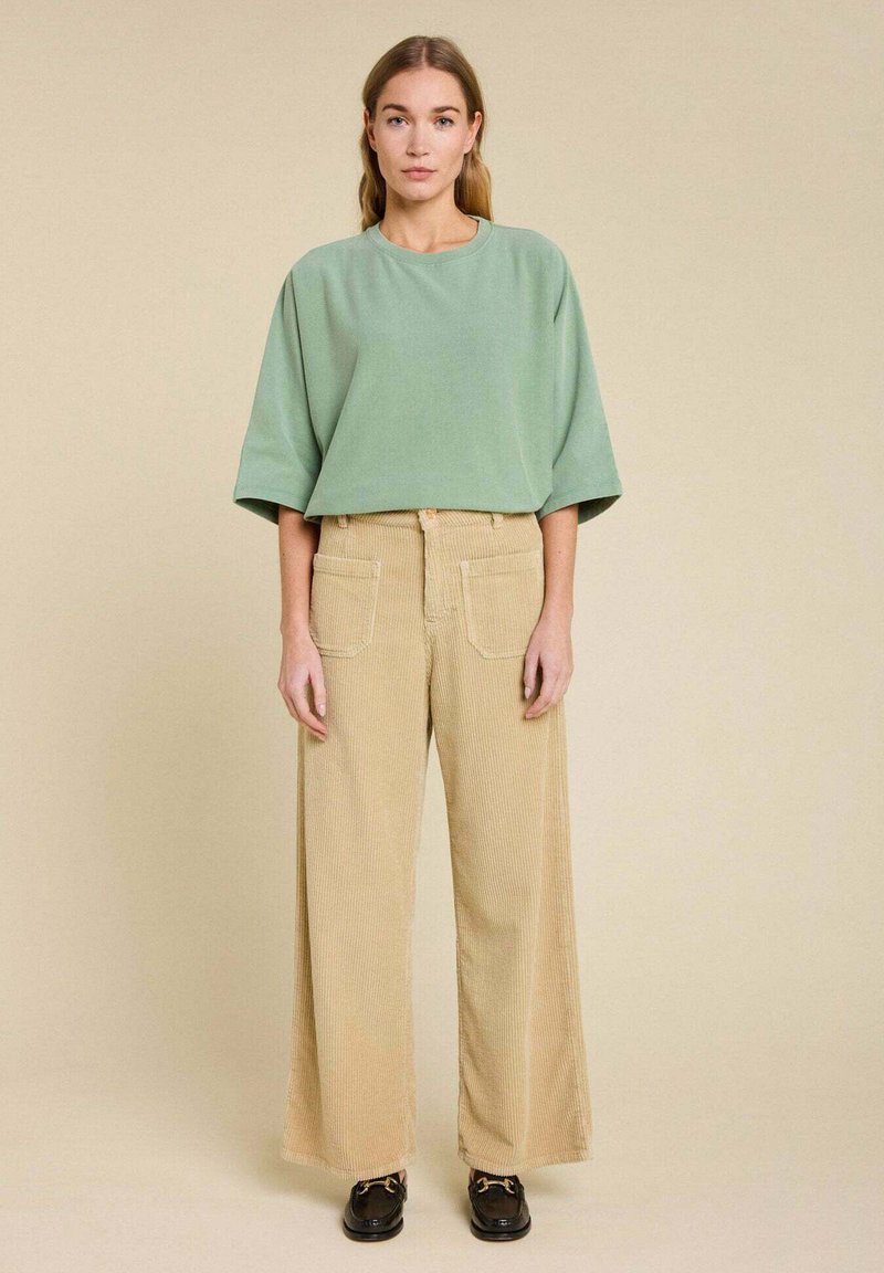 Un haut ample vert menthe à manches courtes associé à un pantalon en velours côtelé large beige avec de grandes poches arrière et des chaussures noires à lacets.